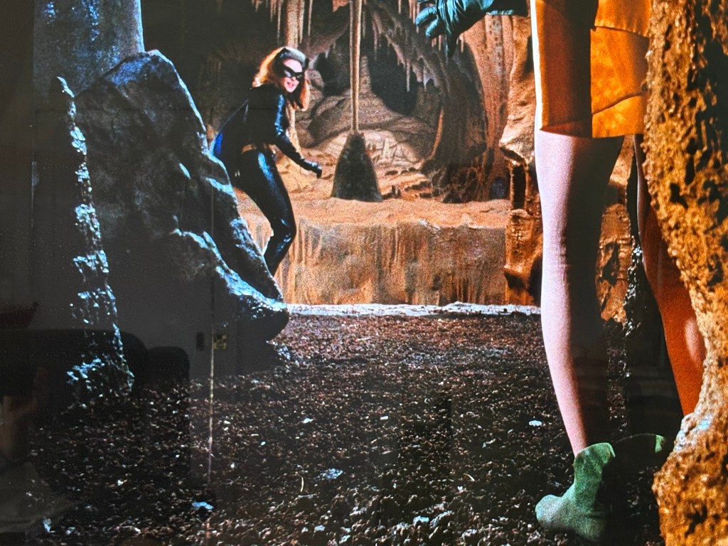 A masked female figure in a black outfit sneaks through a cave-like environment, with rocky formations in the background, while a person in a yellow dress and green shoes stands in the foreground.