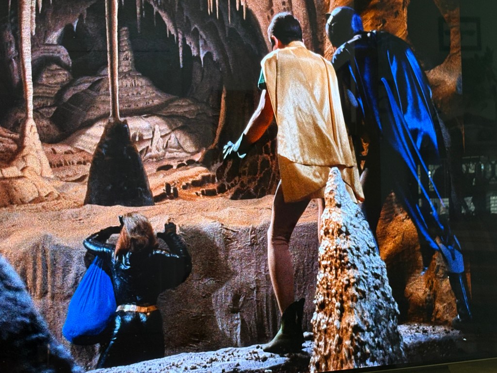 A scene depicting three characters in a cave setting, two of them wearing brightly colored capes and one dressed in dark attire, with the cave's stalactites and formations visible in the background.
