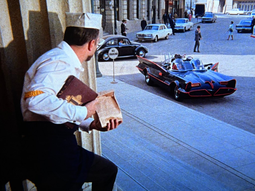 A man in a uniform holding a brown bag and a book looks out from a building entrance as a classic black car with red accents, resembling the Batmobile, drives past on a city street.