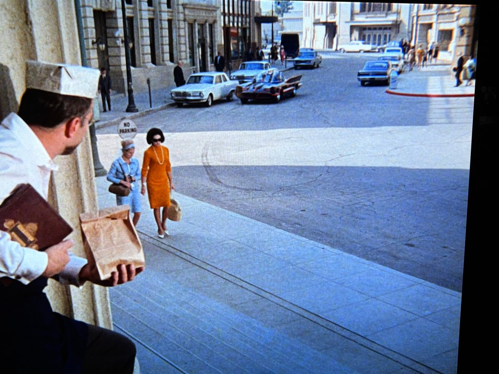 A man in a hat watches two women walking down a street in an urban setting. One woman is dressed in an orange dress while the other is in a light blue outfit. Classic cars are parked along the street, and a 'No Parking' sign is visible.