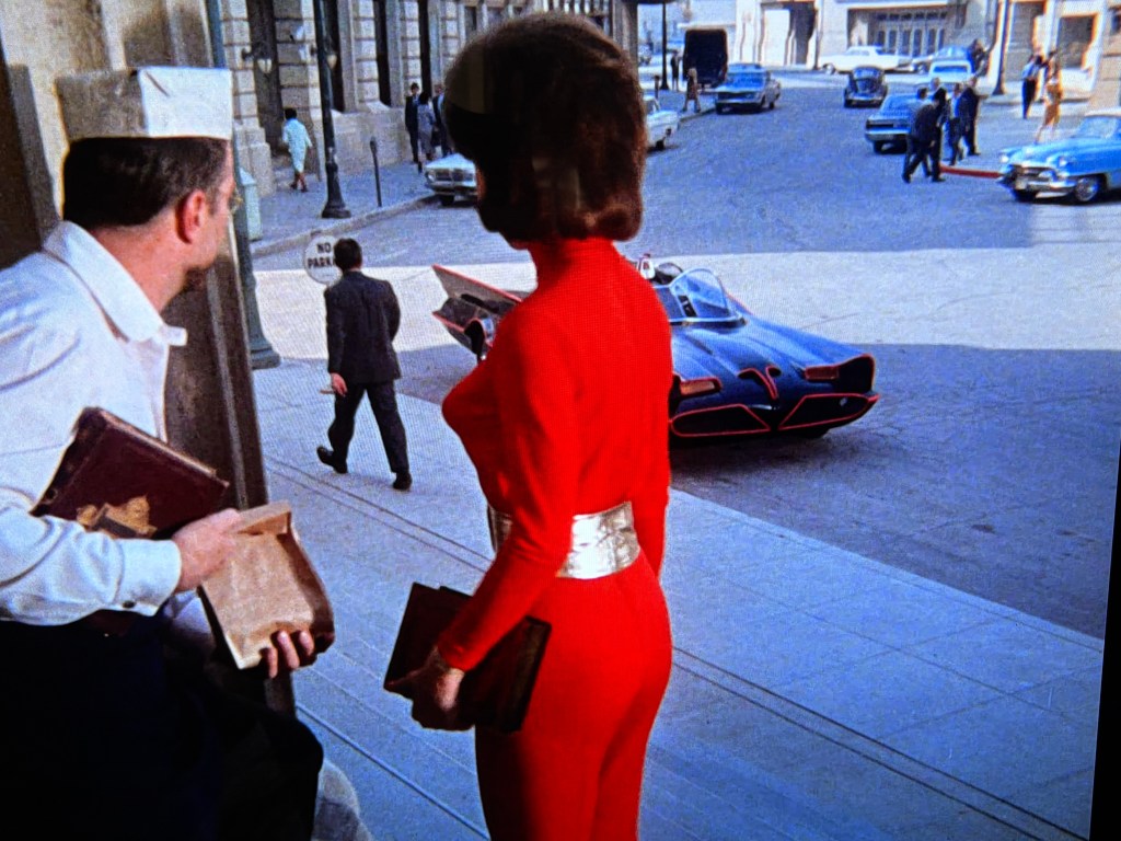 A woman in a red outfit stands on a street, looking away, as a man in a white chef's hat watches her. In the background, a sleek black car is parked, and several people walk down the street lined with buildings.