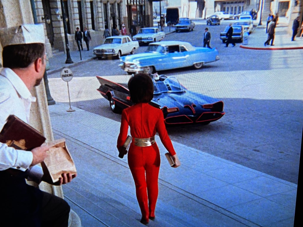 A woman in a red outfit walks towards a black and red iconic car on a city street, while a man in a white uniform looks on from a building entrance.