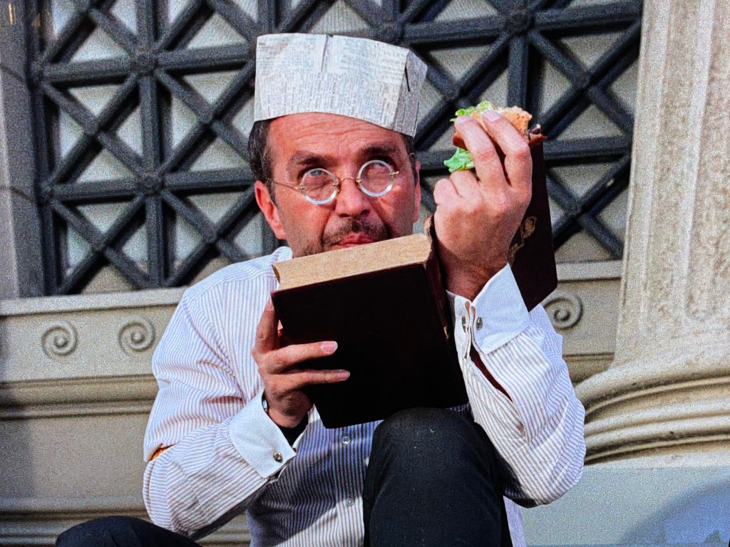 A man wearing a newspaper hat sits while holding a sandwich in one hand and a book in the other, appearing to read and snack simultaneously.