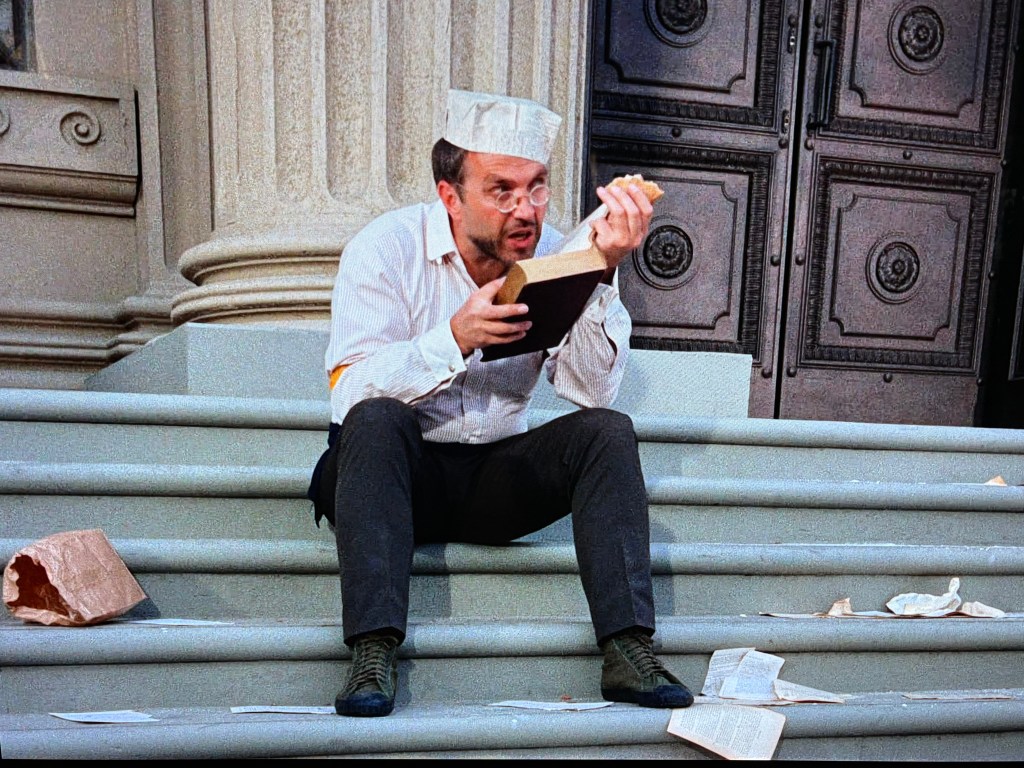 A man wearing a white hat and glasses sits on stone steps, holding a book in one hand and a sandwich in the other, surrounded by scattered papers.