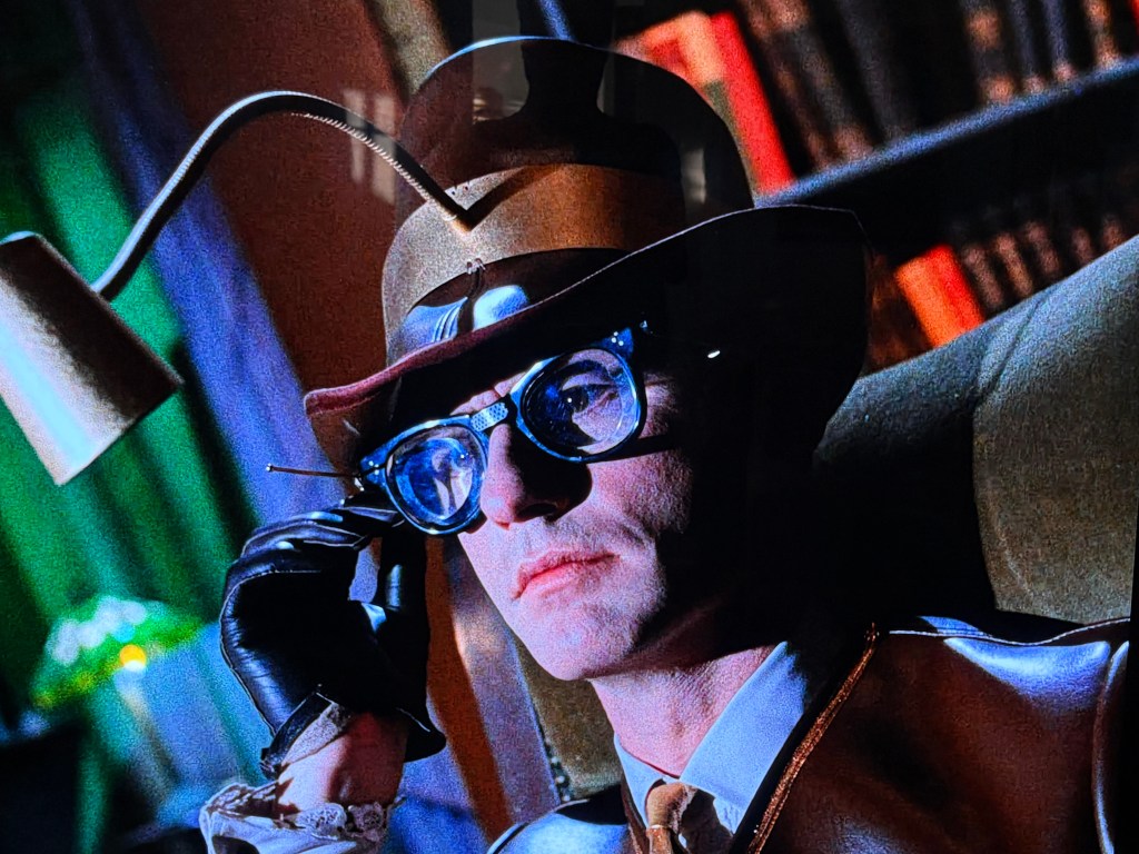 A man with a unique hat and thick glasses sits thoughtfully in a chair, holding a small object to his temple, surrounded by colorful lighting and books.