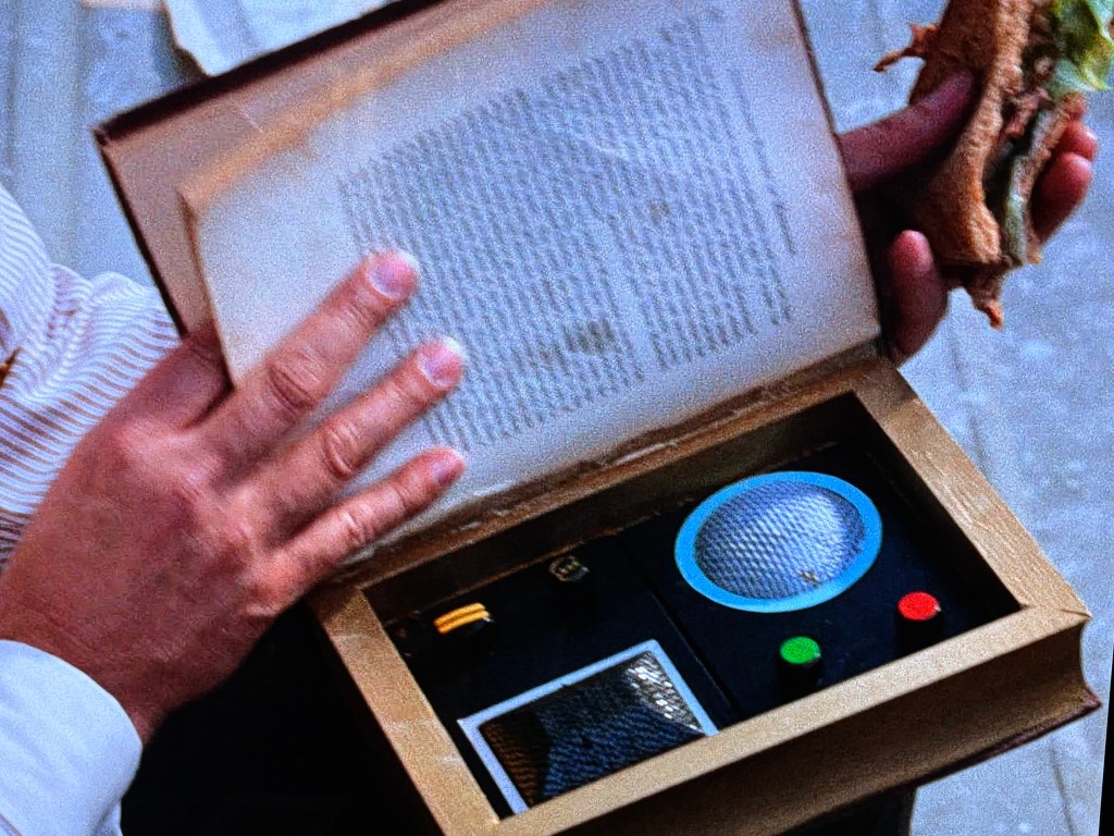 A person holding a sandwich in one hand and a book that contains a hidden gadget with buttons and a speaker in the other hand.