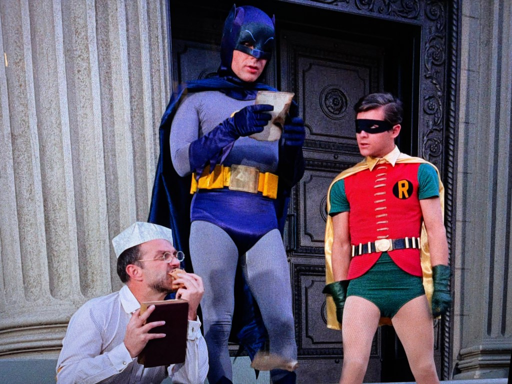 Batman and Robin in superhero costumes, standing in front of a large door, with a man in a paper hat eating a sandwich nearby.