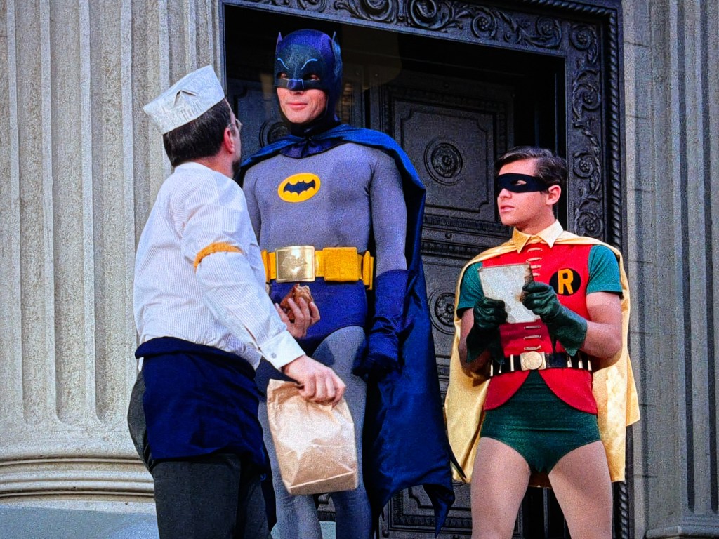 Batman and Robin interacting with a man in a delivery uniform, standing in front of a building entrance.