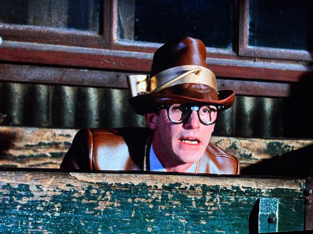 A man with glasses and a brown hat, wearing a brown leather jacket, looks intently from behind a wooden barrier, with a focused expression.