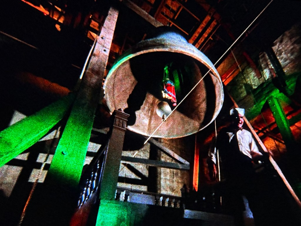 A person stands in a dimly lit space near a large bell, with green and orange lighting illuminating the surroundings. The bell is suspended above, and a rope is attached to it, while wooden structures are visible in the background.
