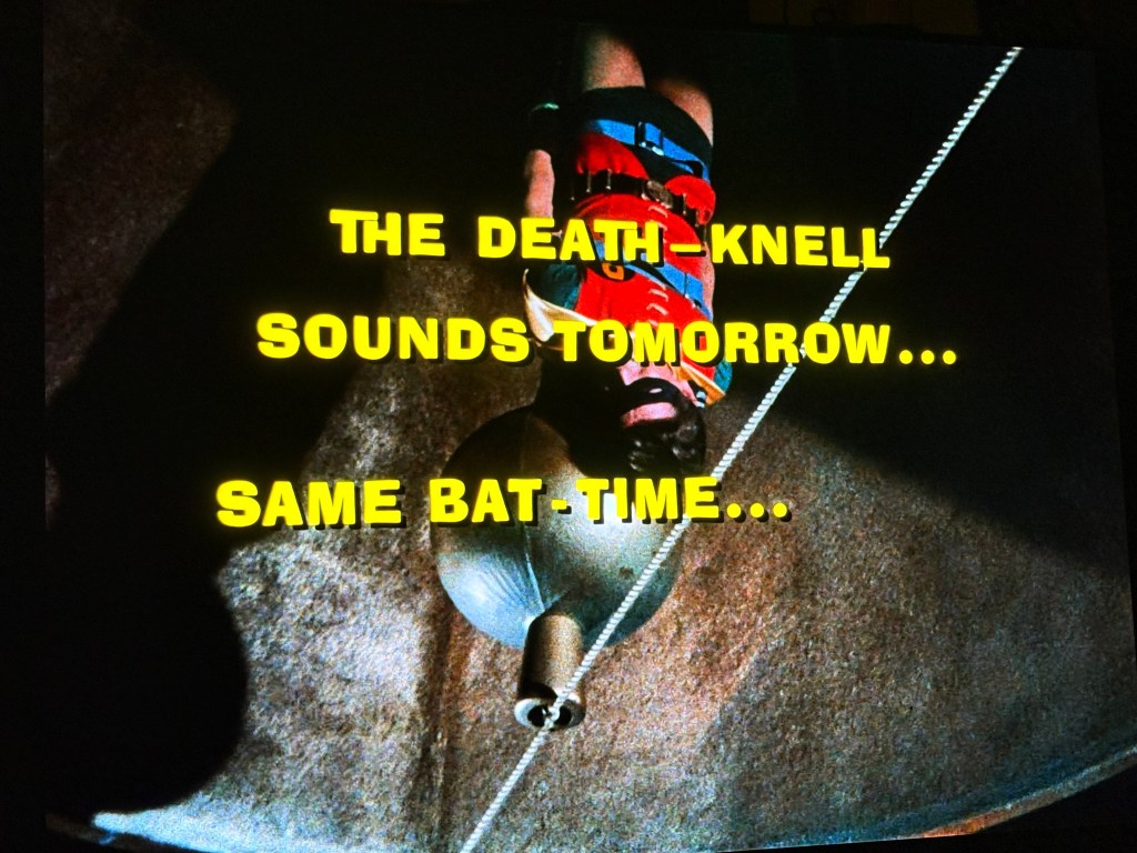 A dramatic scene featuring text that reads 'THE DEATH-KNELL SOUNDS TOMORROW... SAME BAT-TIME...' over an image of a character suspended upside down.