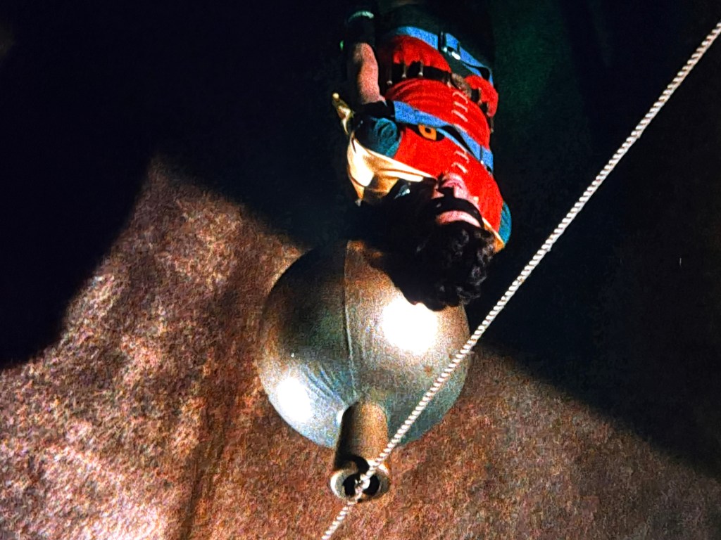 A person in a colorful outfit is suspended above a large metallic sphere, with shadows and a rope visible in the background.