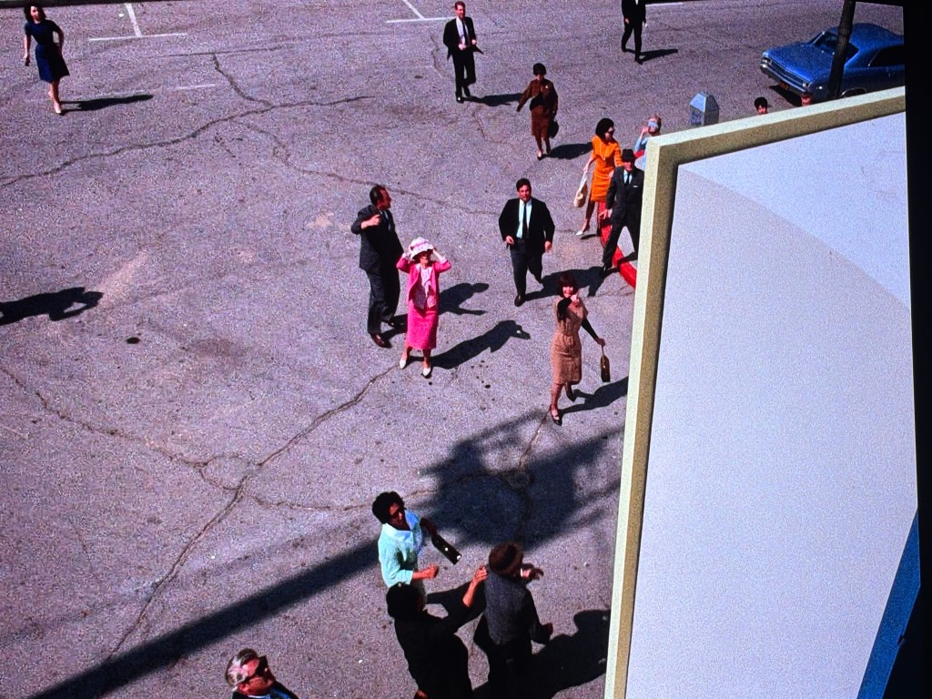 Aerial view of a bustling street with people walking, wearing 1960s attire, including formal suits and dresses. Several individuals appear to be waving or pointing, while cars are parked in the background.