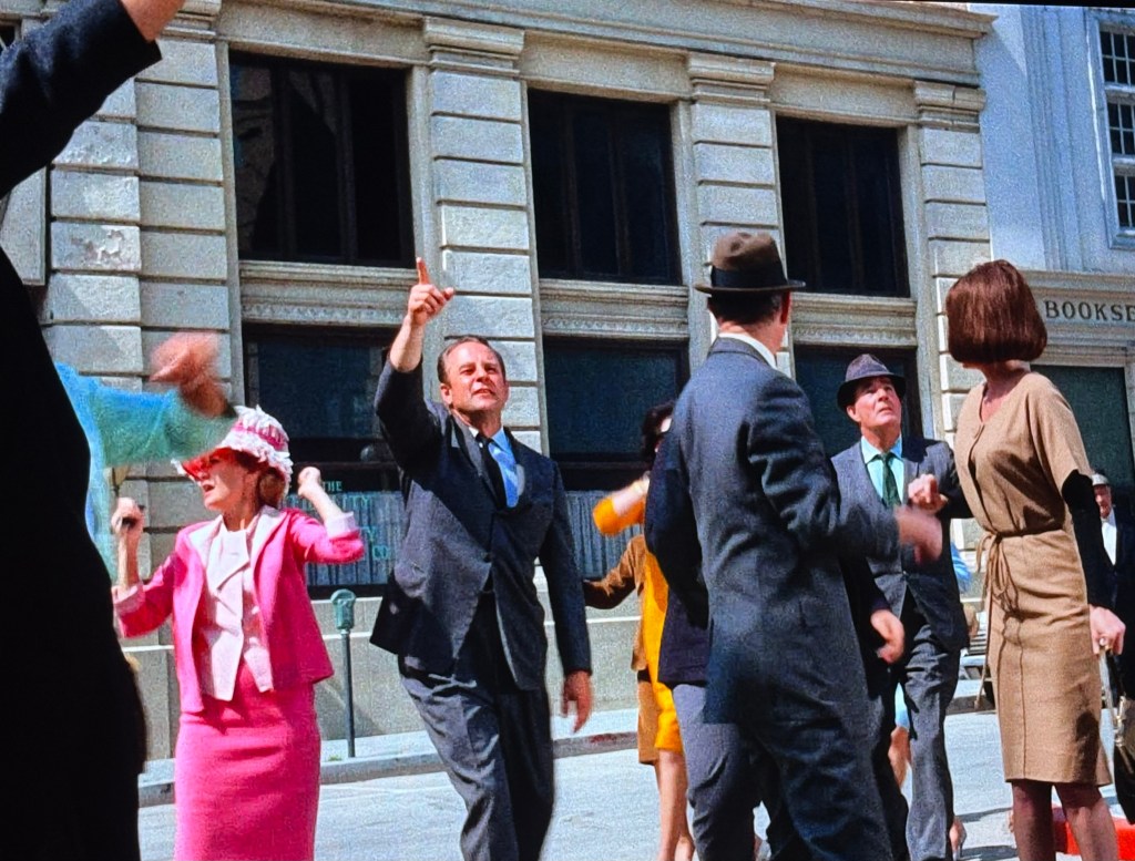 A lively street scene with several people dressed in 1960s fashion, engaging in a spontaneous celebration or dance. A woman in a pink outfit and hat is prominent, while others are smiling and raising their arms in enjoyment. The backdrop features a building with large windows.