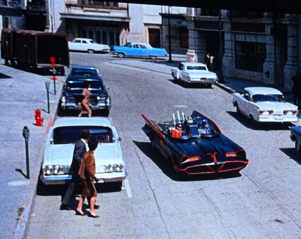 A classic scene featuring a futuristic black car driving through a city street, with several vintage cars parked along the curb. Two pedestrians, a man and a woman, are walking on the sidewalk, while another figure is walking nearby, creating a bustling urban atmosphere.