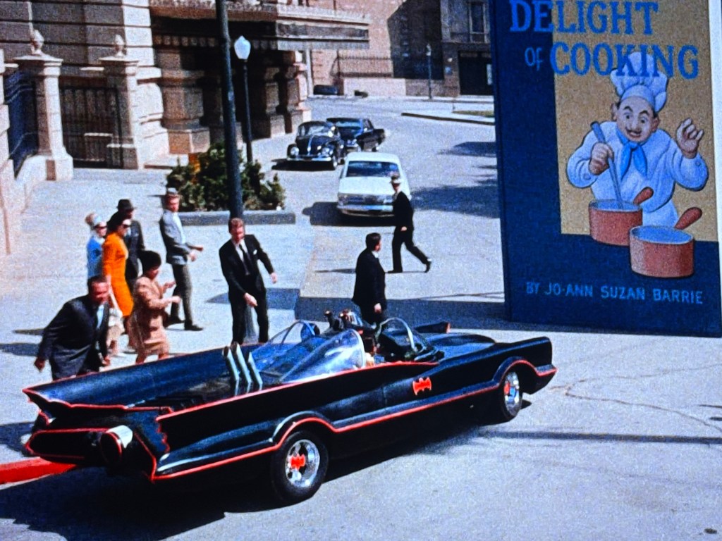 A classic black convertible car, resembling the Batmobile, parked on a city street with people walking around. In the background, a large advertisement for a cookbook titled 'Delight of Cooking' featuring a cartoon chef is visible.