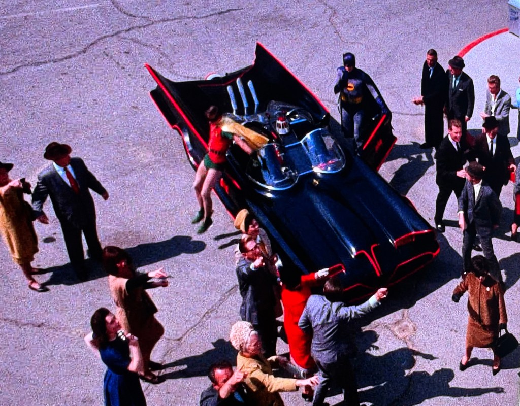 A crowd gathers around a classic black Batmobile with its wings open, featuring Batman and Robin in superhero costumes amidst onlookers in vintage clothing.