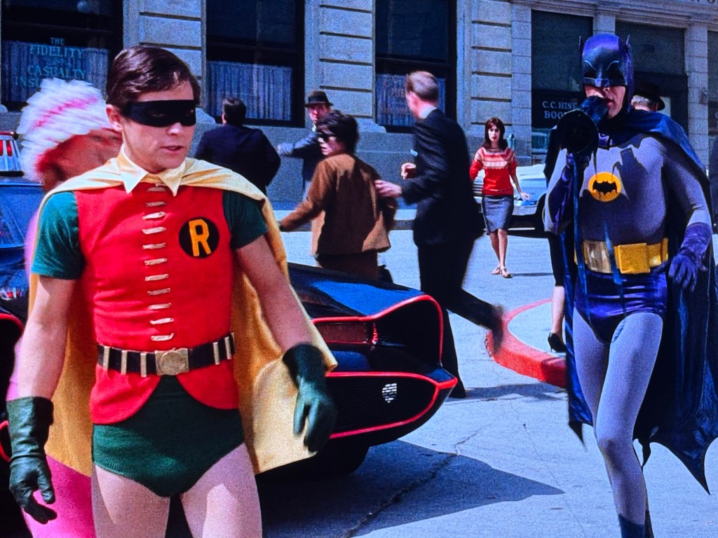 Two actors dressed as Robin and Batman stand in a busy city street, with pedestrians and a parked Batmobile in the background.