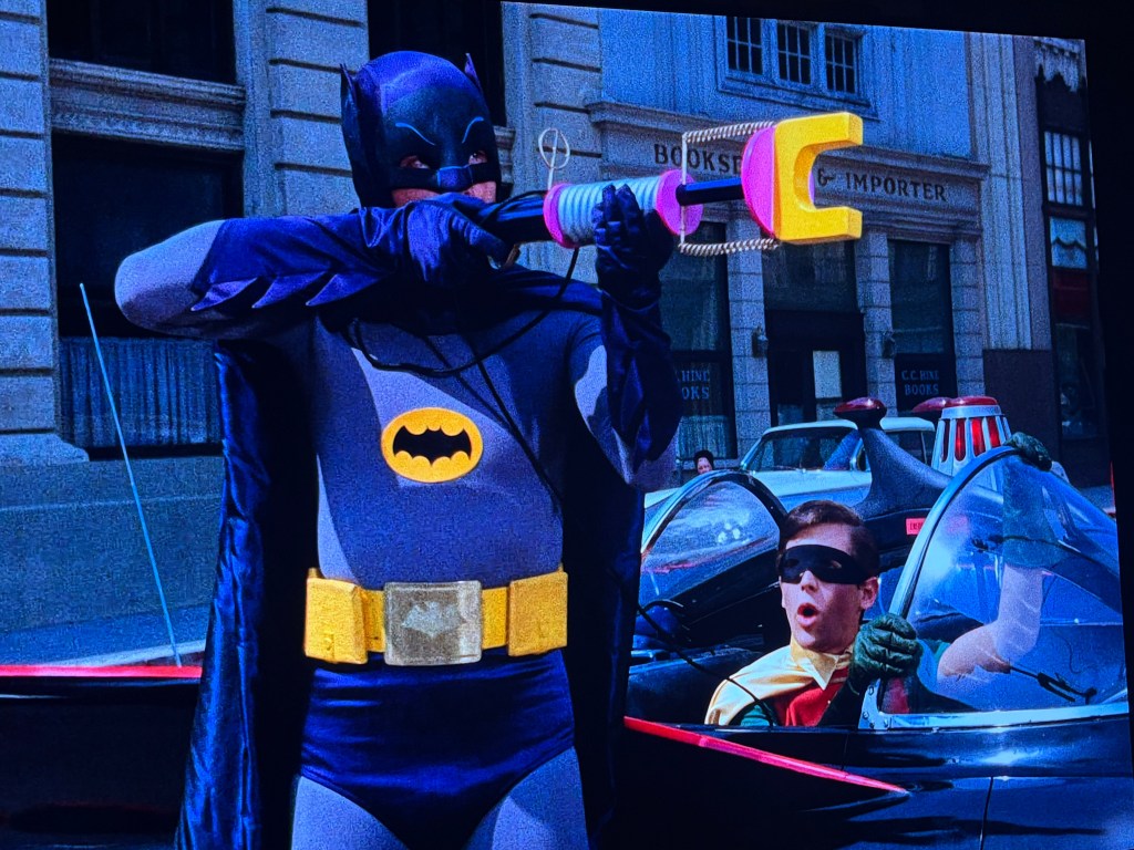 Batman using a gadget from the Batmobile while Robin looks on from the driver's seat.