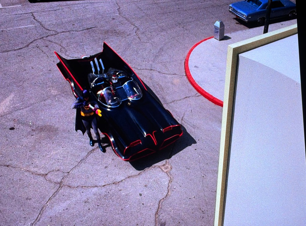 A vintage Batmobile parked on a concrete surface with a character in a Batman costume standing next to it.