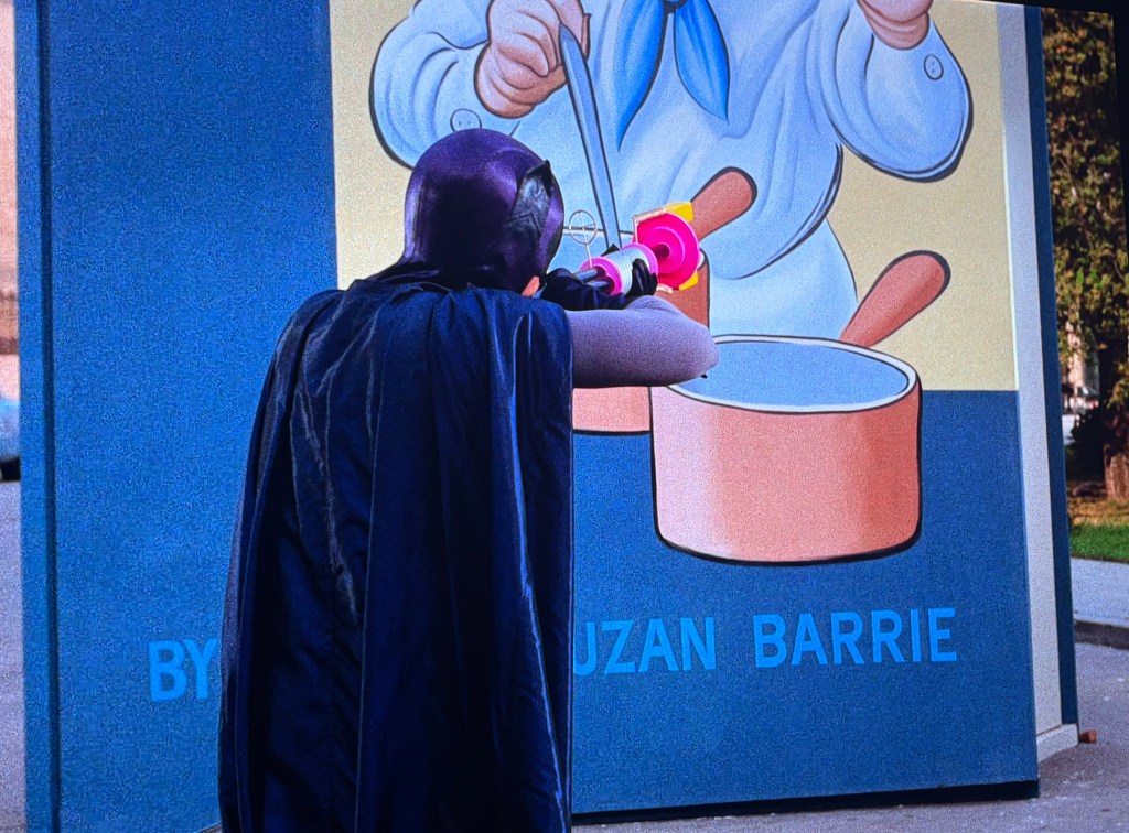 A person wearing a black cape aims a toy gun at a colorful mural depicting a chef stirring a pot, likely in a playful or artistic setting.