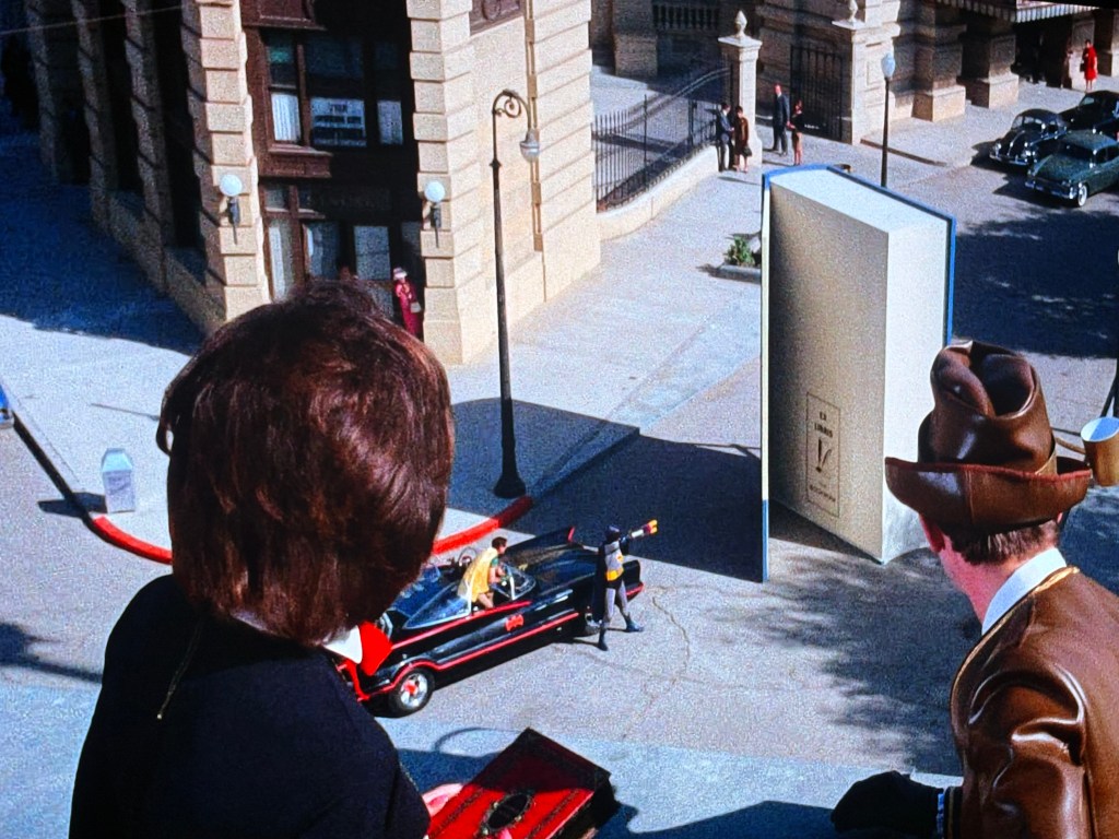 View from above showing two characters observing a street scene with a large book standing upright, while a car and several people are present below.