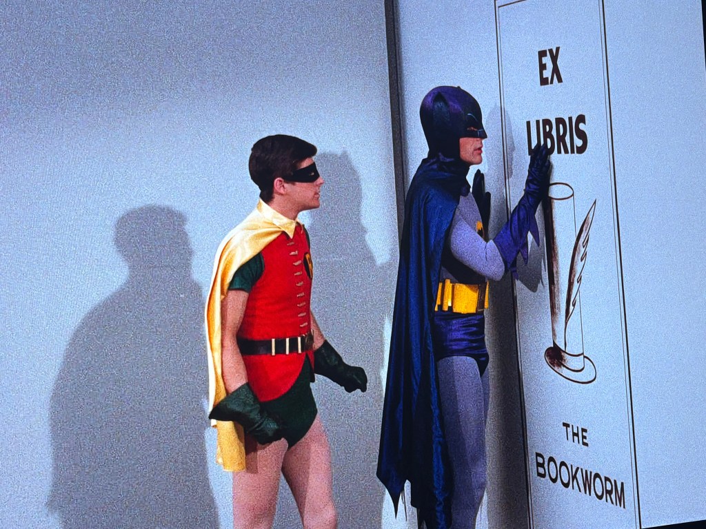 Batman and Robin stand in front of a wall displaying a sign that reads 'Ex Libris The Bookworm'. Batman is touching the sign while Robin looks on.