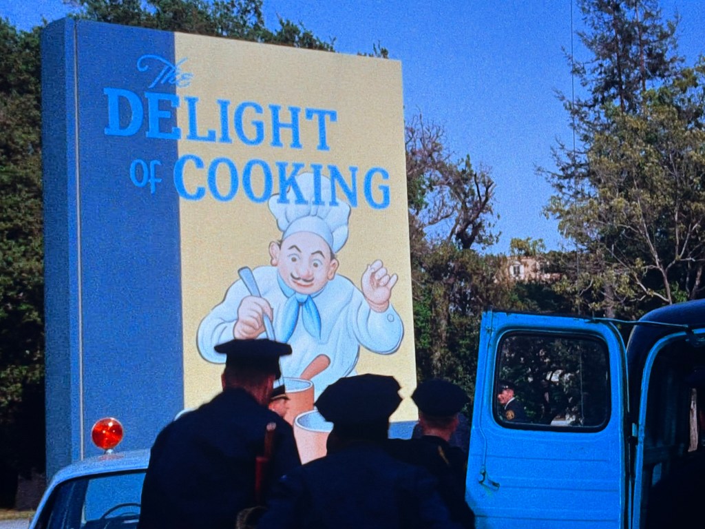 A large colorful sign reading 'The Delight of Cooking' with a cheerful chef illustration, alongside a blue vehicle and several individuals wearing police uniforms.