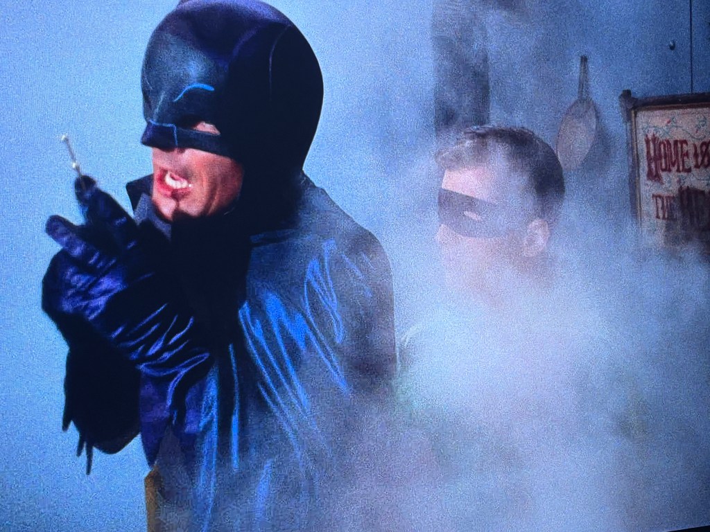A scene featuring two masked characters in a smoky environment, one dressed in a dark cape and cowl, holding a device, while the other, wearing a green outfit and a mask, stands closely behind.