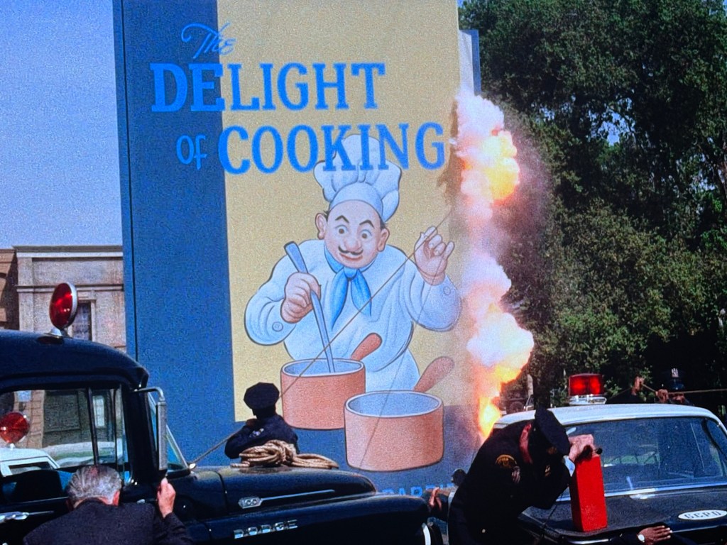 A colorful billboard depicting a cheerful chef with pots, titled 'The Delight of Cooking,' alongside an explosion and emergency vehicles responding.