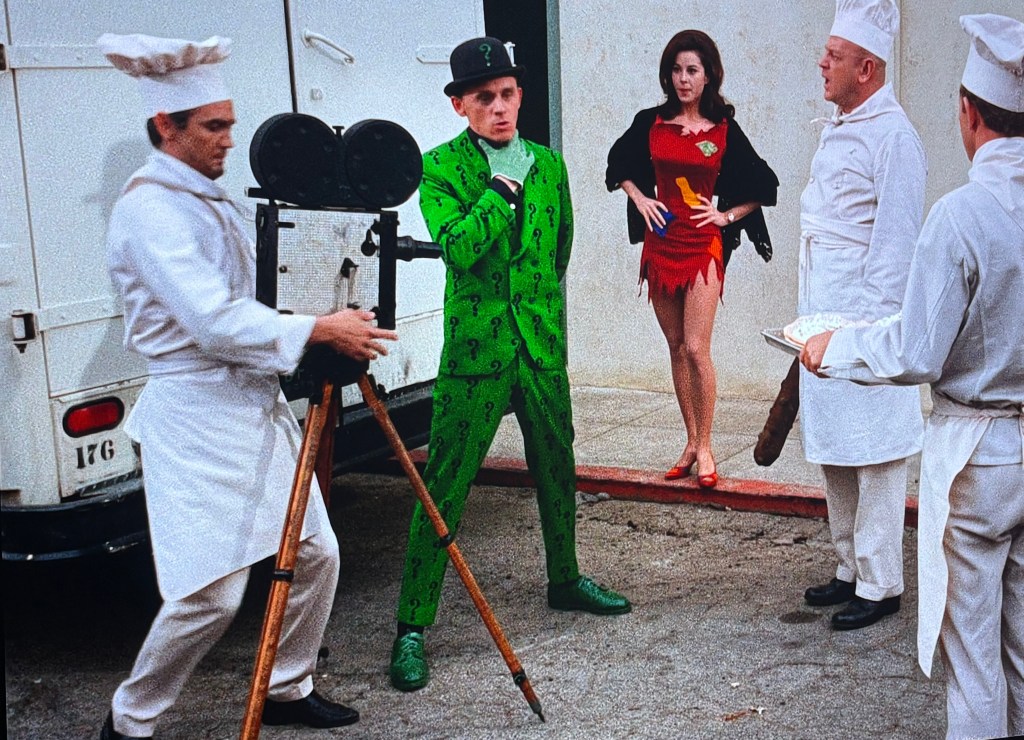 A scene with a man dressed in a green suit with question marks, holding an umbrella and posing in front of a camera. Nearby, a woman in a red and yellow dress stands with her hands on her hips. Two chefs in white uniforms observe, one holding a plate.