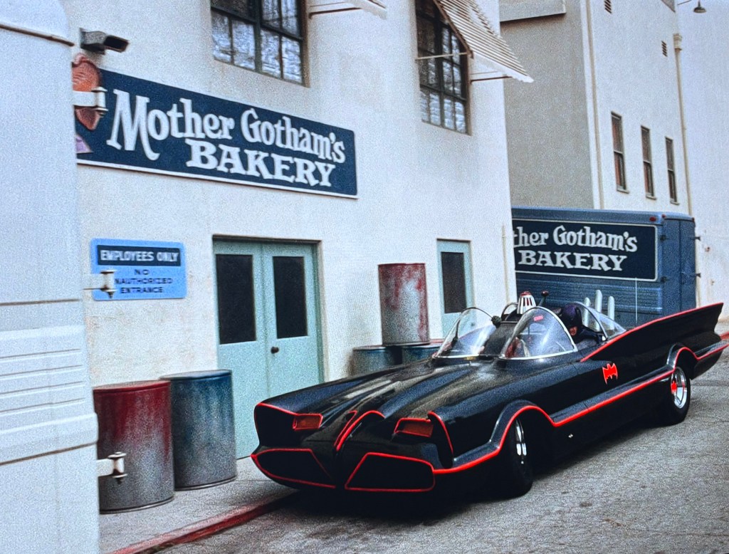 A black and red Batmobile parked outside 'Mother Gotham's Bakery' with a blue sign indicating 'Employees Only' nearby.