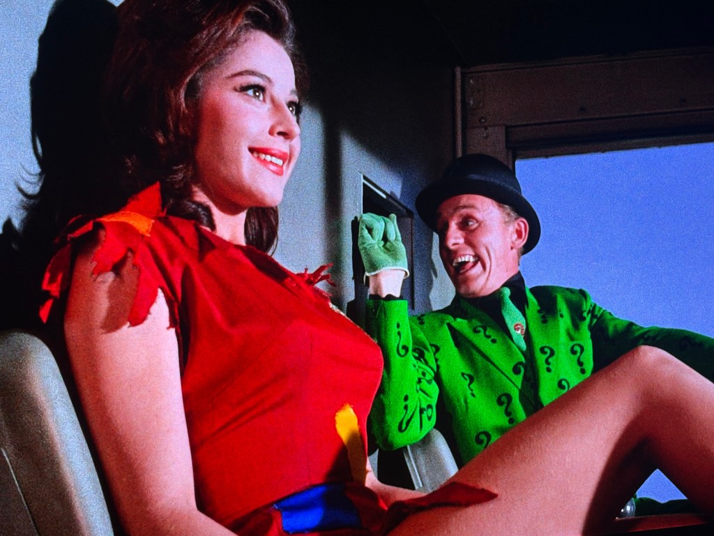 A woman in a bright red dress smiles while sitting beside a man in a green suit with question marks, who appears to be animated and cheerful.