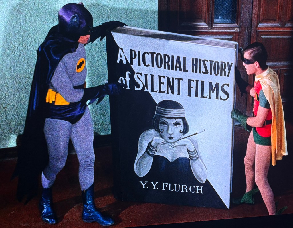 Batman and Robin stand next to a large book titled 'A Pictorial History of Silent Films' by Y.Y. Flurch, with an illustration of a woman holding a conductor's baton.