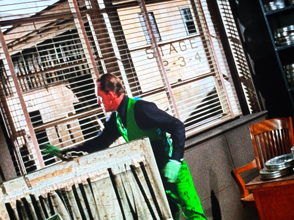 A man in a green outfit is cleaning a dusty surface in a room with large windows and a sign indicating 'STAGE 2-3-4' in the background.