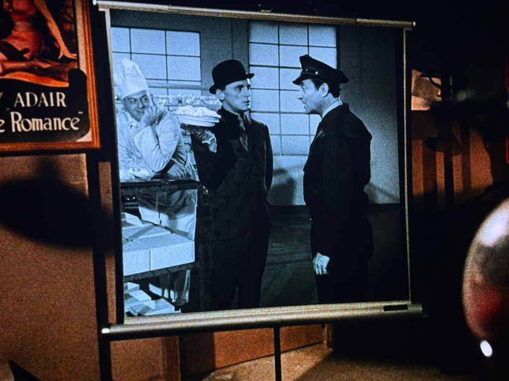 A scene from a classic film shows two men in conversation in front of a large projection screen, with one man dressed in a police uniform and the other in a hat and formal attire. In the background, a chef is watching from behind a counter.
