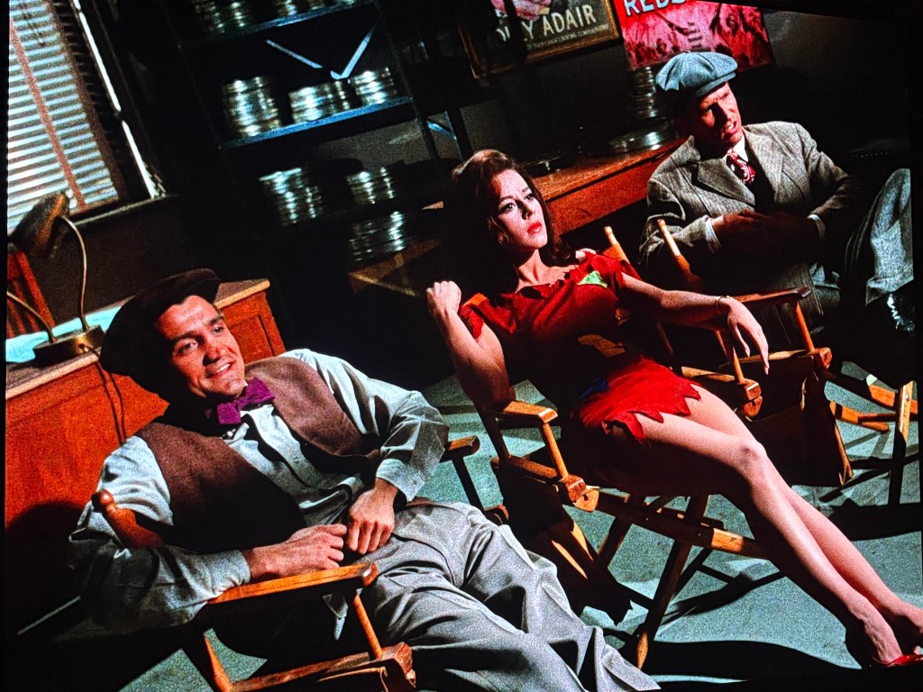 Three individuals sitting on director's chairs in a film studio, with film reels and posters visible in the background. The woman is wearing a red dress, while the men are in formal attire.
