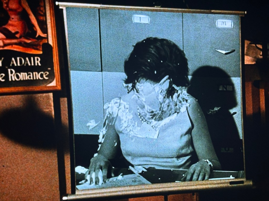 A woman with her face covered in what appears to be cream or pie, sitting at a desk in front of a wall of filing cabinets.