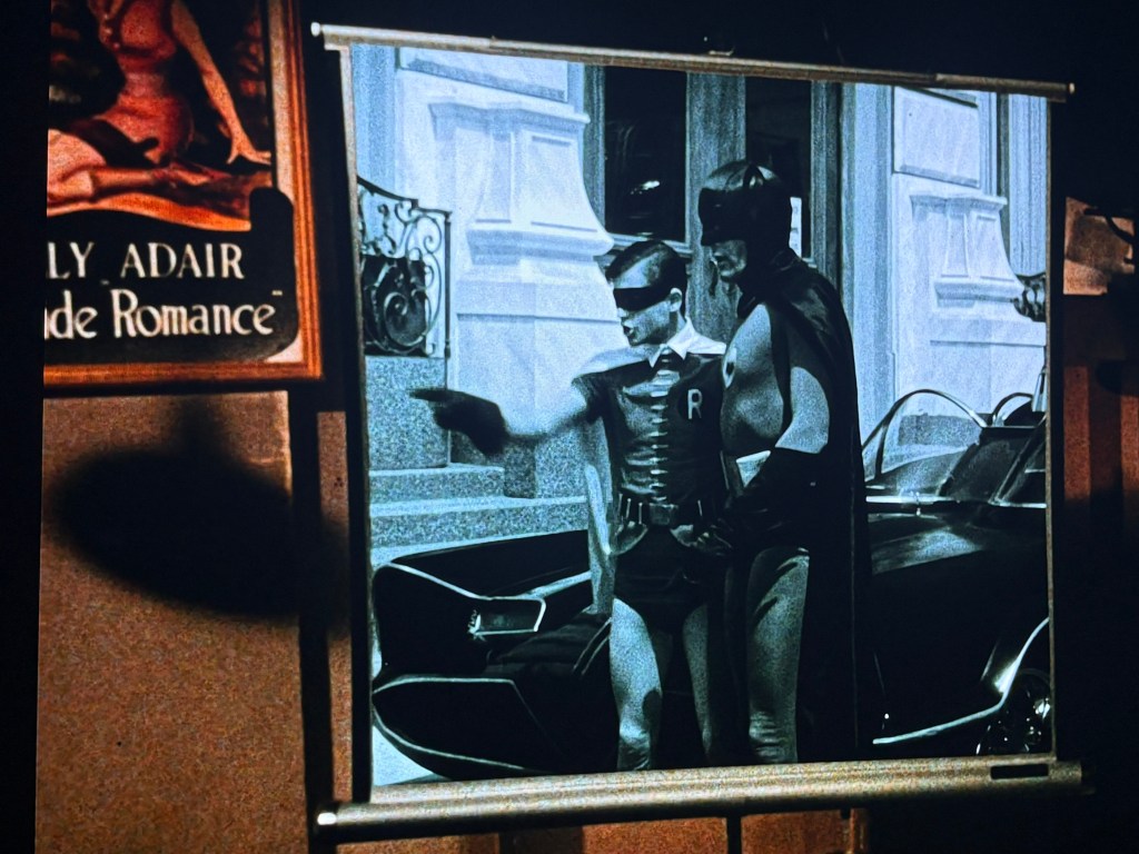 A black and white image featuring a movie scene of a superhero duo, Batman and Robin, standing next to a classic car. Robin is pointing while Batman looks on, both in their iconic costumes.