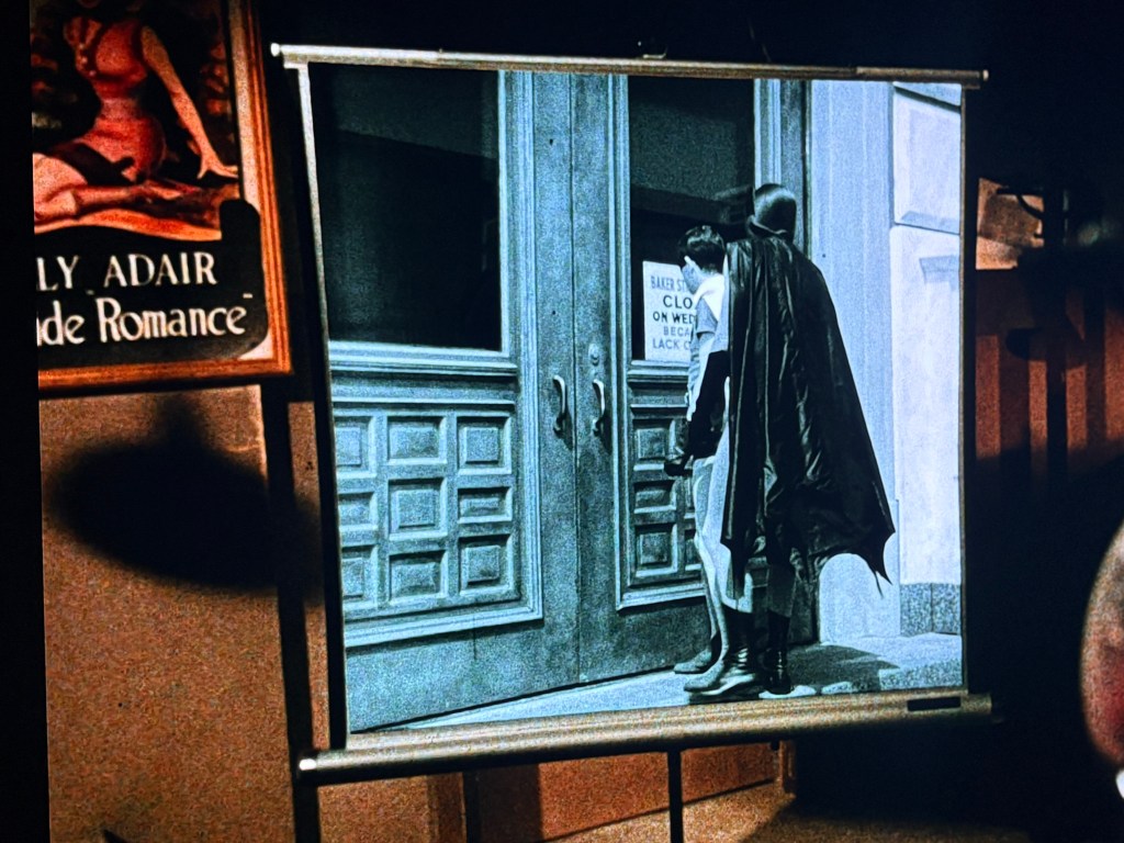A black and white film scene featuring a superhero in a cape and a child standing in front of a movie theater entrance, with a sign stating the theater is closed.