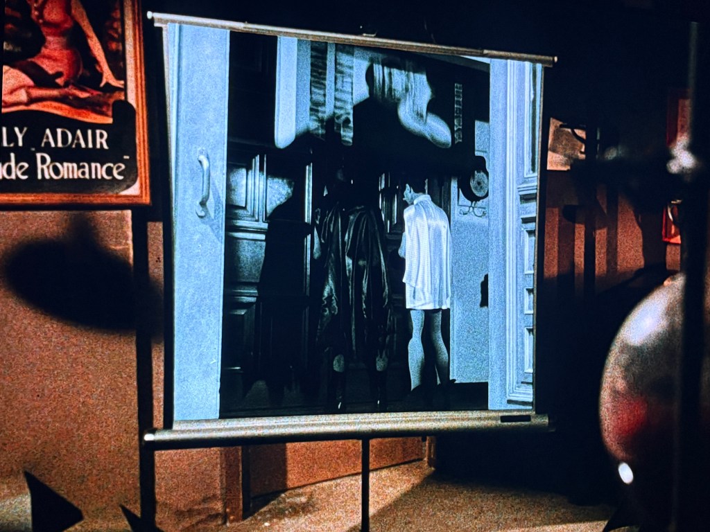 A vintage projector screen displaying a black and white image of two figures indoors, with a contrasting colorful background featuring an advertisement for 'Lily Adair, Nude Romance.'