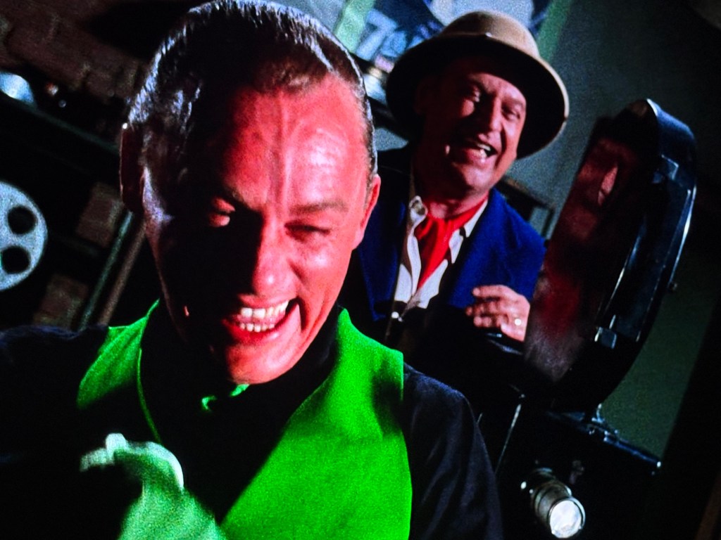 Two men smiling and laughing, one wearing a bright green outfit and the other in a blue suit and hat, with a film projector in the background.