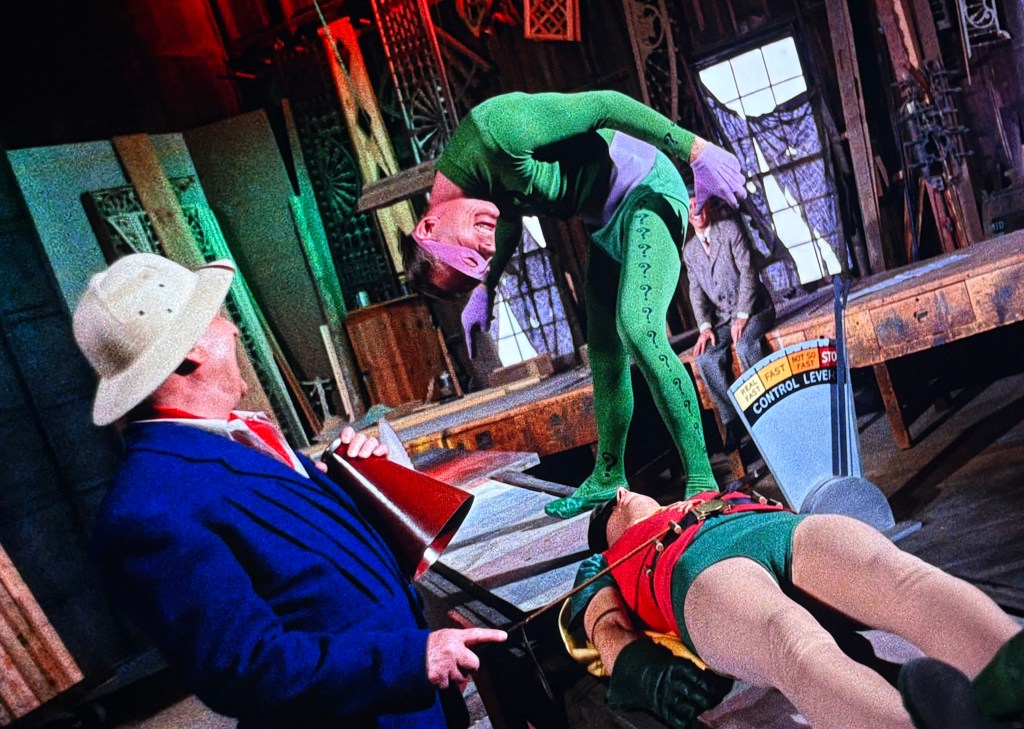 A scene depicting a colorful villain, dressed in a green costume with a question mark motif, looming over a tied-up hero in a red and green outfit. In the background, a man in a white hat is holding a megaphone, while another figure looks on, all set in a dimly lit room filled with various props.