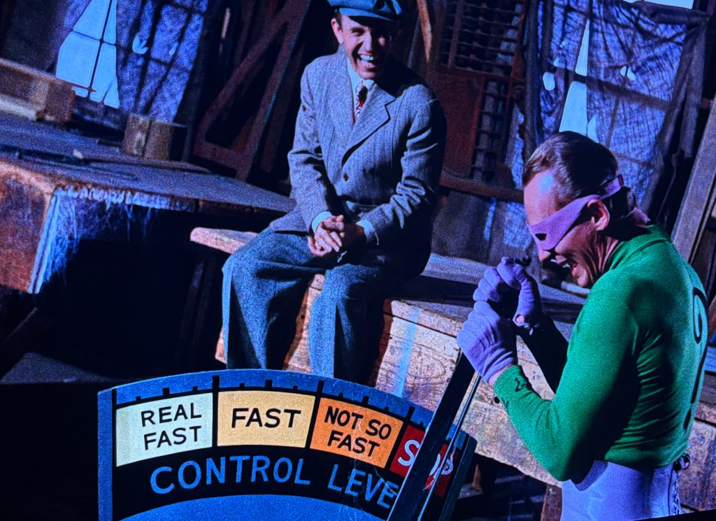 A scene featuring two characters, one in a suit and hat laughing, and another wearing a green superhero costume holding a panel labeled 'CONTROL LEVELS' with indicators for speed.