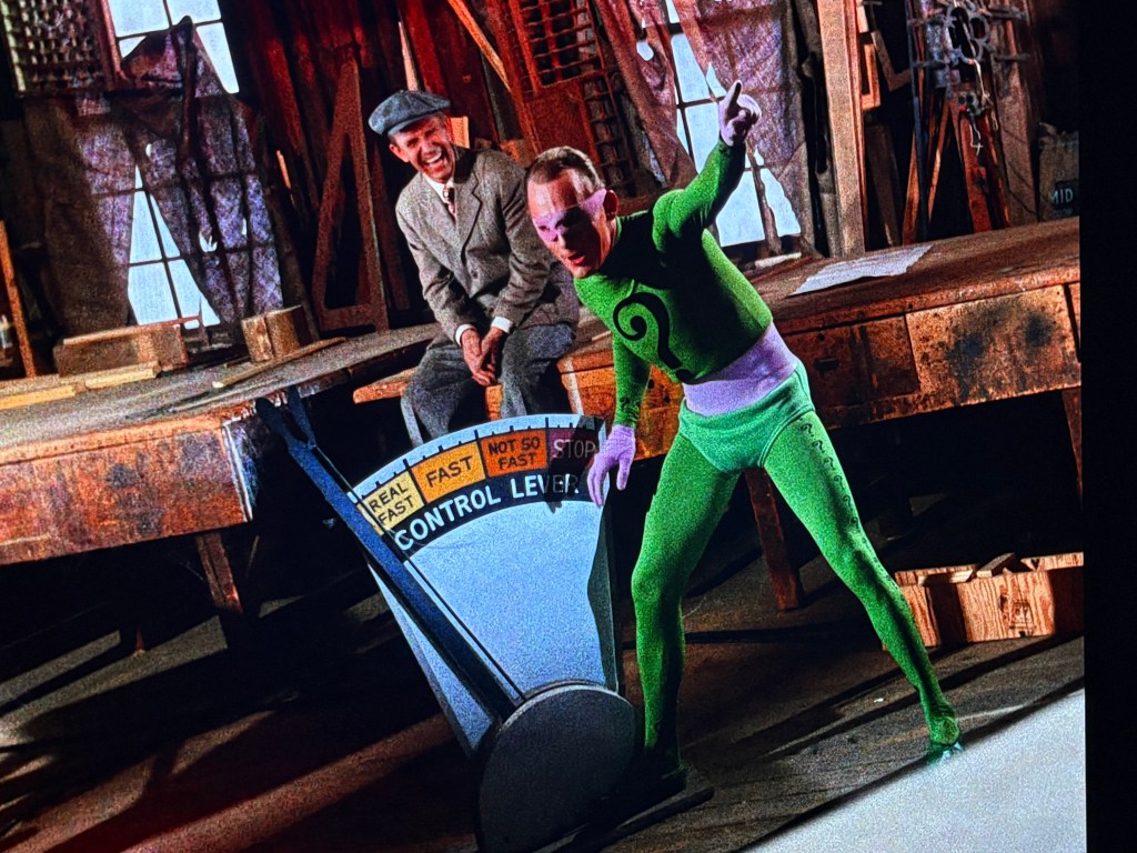 A man in a green costume with purple accents gestures dramatically in front of a large control lever marked 'CONTROL LEVEL.' Another man wearing a gray suit and cap laughs in the background, inside a rustic wooden room.
