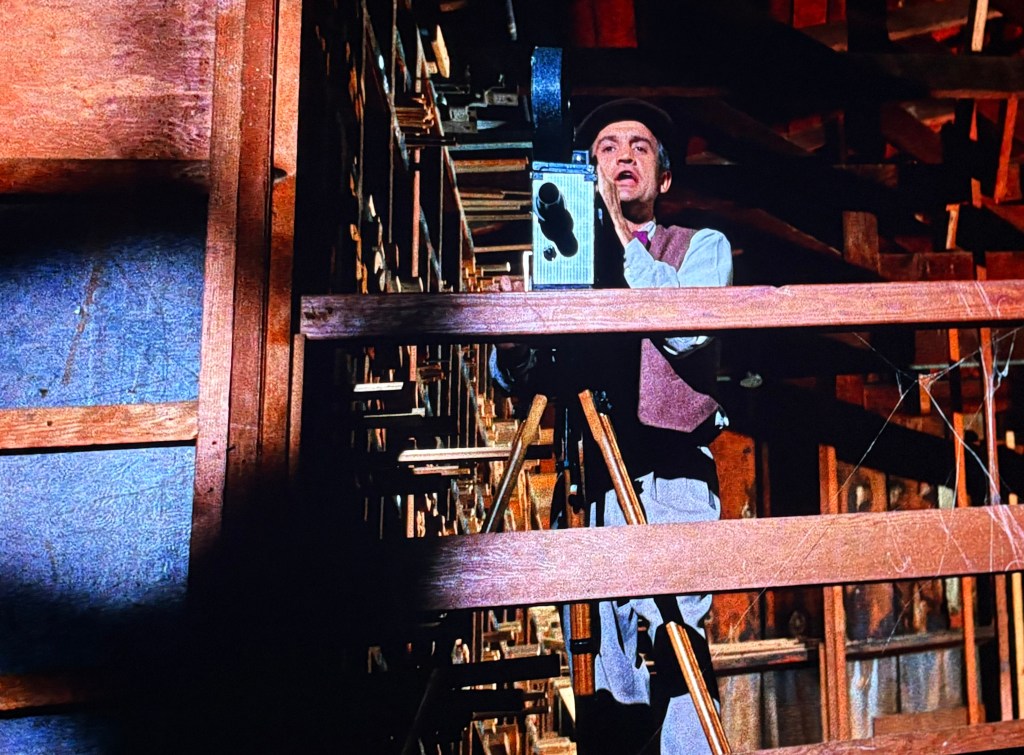 A man using an old-fashioned camera on a wooden platform surrounded by a rustic interior, with beams and supports visible in the background.