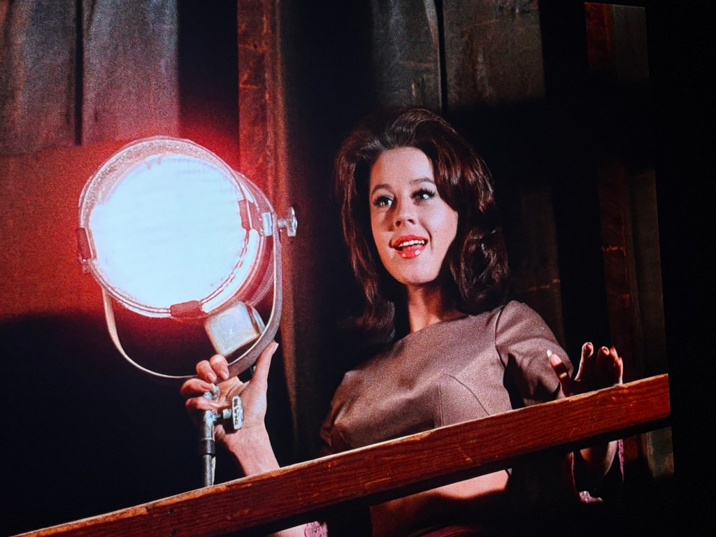 A woman with dark, styled hair is holding a spotlight while smiling, set against a dimly lit background.