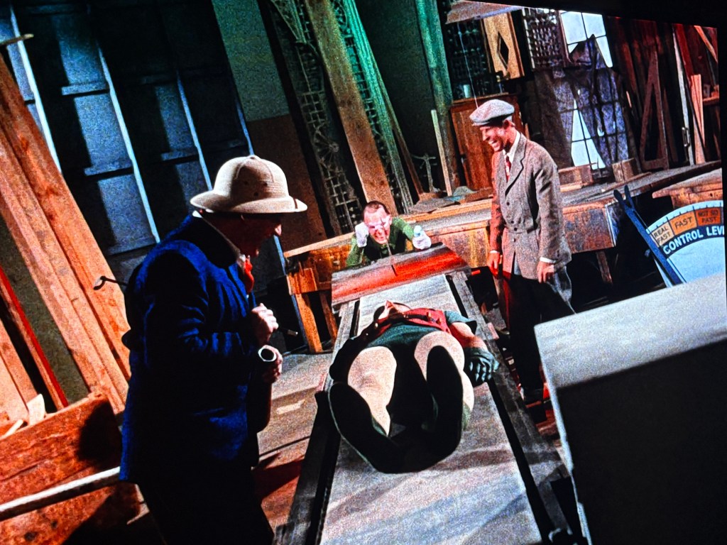 Four men in a cluttered workshop, with one man lying on a table while others appear to be inspecting him. The setting features wooden materials and tools, suggesting a stage or production environment.