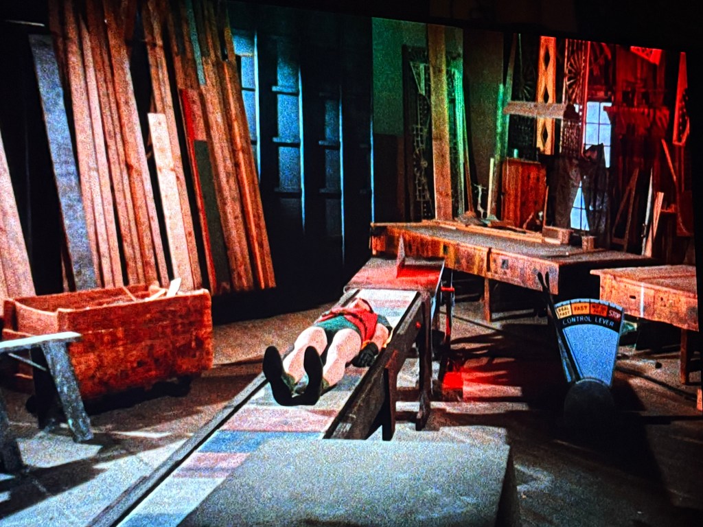 A dimly lit workshop with wooden beams and tools scattered around. A person lies on a wooden plank surface near a table, seemingly in an inactive pose. The scene has colorful lighting effects, creating a dramatic atmosphere.