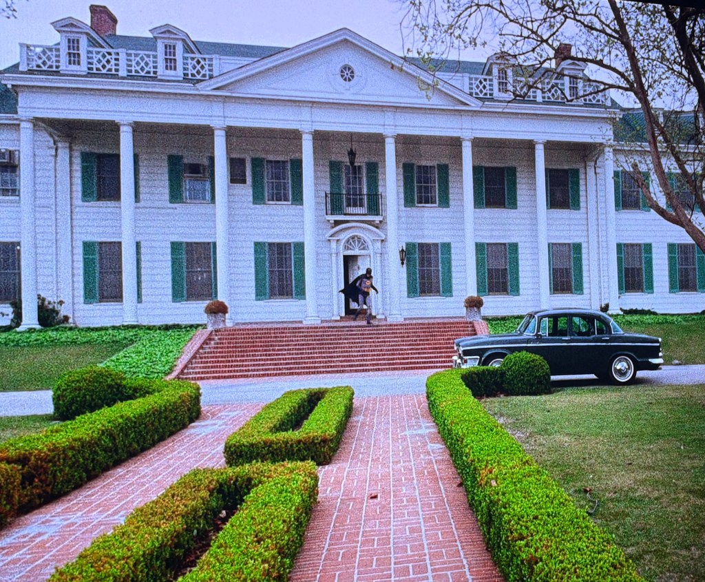 A grand white mansion with green shutters, featuring a large staircase leading to the entrance. A person in a dark cape is seen on the steps, while a vintage car is parked in front amidst neatly trimmed hedges.